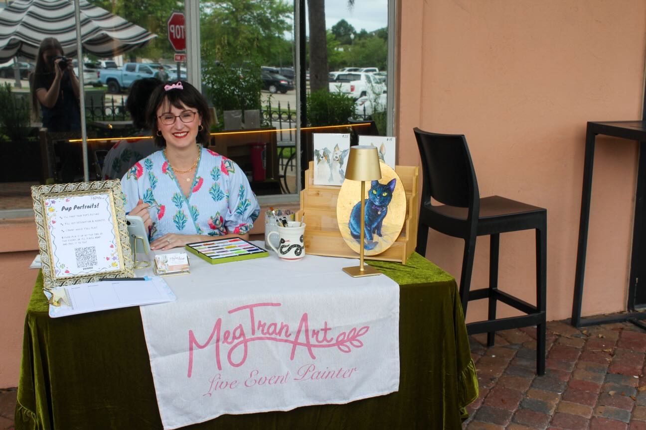 Woman sitting at a table with art supplies and a sign reading 'Meg Tran Art' in an outdoor setting.